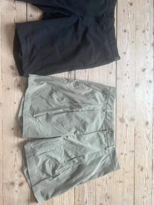 Två par shorts, svarta och beige - Säljer två par shorts, ett par svarta och ett par beige. De svarta har en enkel design med elastisk midja, medan de beige har klassisk passform med bälteshällor och knappstängning. Perfekta för sommaren och lätta att matcha till olika outfits.