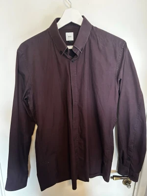 Skjorta från bläck  - Säljer en stilren mörklila skjorta från bläck med klassisk krage och knappar framtill. Skjortan är långärmad och har en clean look utan mönster, perfekt för dig som gillar minimalistisk stil. Pris kan diskuteras🙌