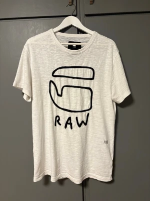 Vit t-shirt från G-Star RAW - Vit t-shirt från G-Star RAW med svart grafiskt tryck och texten RAW på framsidan. Klassisk rund hals och korta ärmar. Tillverkad i mjuk bomull för en skön känsla och avslappnad passform.
