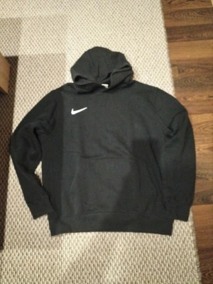 Svart Nike hoodie med huva - Säljer en svart hoodie från Nike med klassisk vit swoosh på bröstet. Hoodien har en stor huva och en praktisk magficka. Materialet är mjukt och bekvämt, perfekt för chill dagar eller när du vill ha en sportig look.