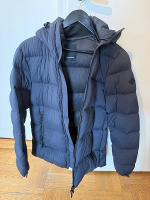 Svart dunjacka från J.Lindeberg - Snygg svart dunjacka från J.Lindeberg med huva och dragkedja framtill. Jackan har två sidofickor med dragkedja och en diskret logga på ärmen. Perfekt för kalla vinterdagar och riktigt skön att bära tack vare det vadderade materialet.