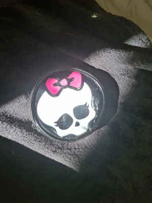 Monster High rund plånbok svart/vit - Säljer en rund plånbok från Monster High i svart och vitt konstläder. Framsidan har ett tryck av en vit döskalle med rosa rosett och svarta detaljer. Baksidan är helsvart. Perfekt för småsaker eller mynt och stängs med dragkedja.