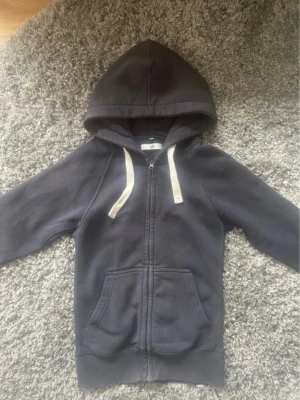 RusseMerch  - Mörkblå hoodie med dragkedja från Russemerch. Klassisk modell med stor huva, vita dragsnören och två rymliga fickor framtill. Tillverkad i mjuk bomullsblandning som är skön att ha på sig. Perfekt för en avslappnad och stilren look. Lite urtvättad men fin 