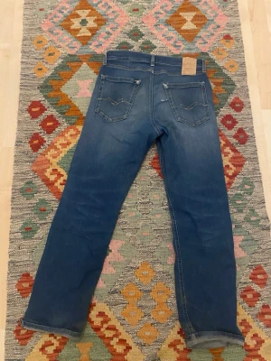 Blå raka jeans från Nudie Jeans - Säljer ett par klassiska blå jeans från Nudie Jeans med rak passform och fem fickor. Jeansen har snygga slitningar och kontrastsömmar, samt Nudie-loggan på bakfickan. Perfekta för en avslappnad och stilren look.