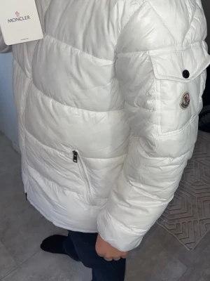 Vit dunjacka från Moncler - Snygg vit dunjacka från Moncler med klassisk logga på ärmen och dragkedjeförsedda fickor. Jackan har quiltad design och innerfoder med serietidningsmönster. Perfekt för kalla vinterdagar och riktigt stilren look.