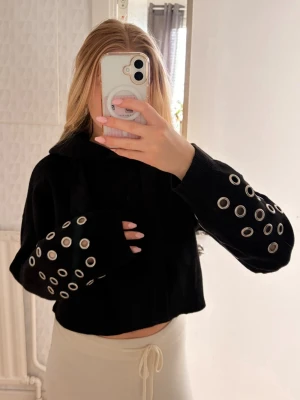 Svart stickad hoodie med detaljer - Säljer en svart stickad cropped hoodie med huva och stora metallringar som detaljer längs ärmarna. Hoodien har långa ärmar och en avslappnad passform. Materialet känns mjukt och stretchigt