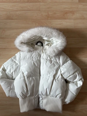 Vit pufferjacka  - Snygg vit pufferjacka med stor huva kantad av fluffig fuskpäls. Jackan har breda ribbade muddar vid ärmslut och nederkant, samt dragkedja framtill. Perfekt för kalla vinterdagar och ger en trendig look.