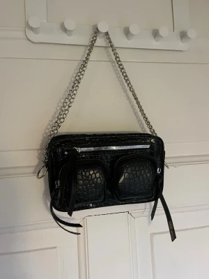 Svart axelväska med kedja - Snygg svart axelväska i croco-mönstrat skinnimitation med två framfickor och silverfärgad kedjerem. Väskan har dragkedjor och coola detaljer som ger en edgy vibe. Perfekt för dig som vill ha en trendig och praktisk väska till vardagen.