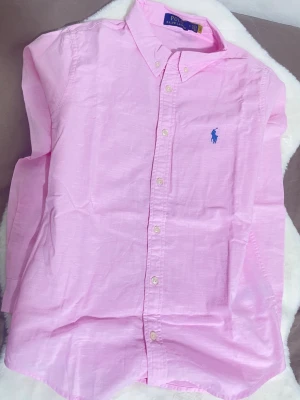 Rosa skjorta från Polo Ralph Lauren - Säljer en klassisk rosa skjorta från Polo Ralph Lauren med button-down krage och blå broderad logga på bröstet. Skjortan har långa ärmar och knäppning framtill. Den är helt ny och i bästa skick! Perfekt för sommaren ☀️Hör av dig vid intresse och mer info🥂.                 Skick[10/10], storlek[M], pris[549].           
