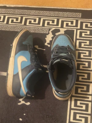 Nike Dunk Low blå och marin sneakers - Nike Dunk Low sneakers i blått och marinblått läder med vit swoosh på sidan. Klassisk låg siluett med perforerad tåbox och matchande blå skosnören. Yttersulan är vit med mörkare detaljer. Perfekt för dig som gillar streetwear och snygga färgkombos.