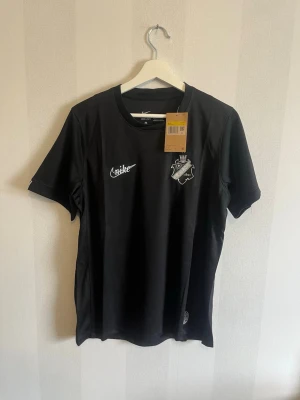 Svart AIK Nike Dri-FIT t-shirt - Snygg svart t-shirt från Nike med AIK:s klubbmärke på bröstet och vit Nike-logga. Tillverkad i Dri-FIT-material som andas, perfekt för träning eller match. Rund halsringning och korta ärmar. En stilren supportertröja för AIK-fans.
