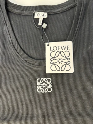 Mörkgrå ribbad linnetopp  - Snygg mörkgrå ribbad linnetopp  med rund halsringning och broderad logga framtill i vitt. Perfekt att styla till jeans eller kjol för en clean look. Materialet känns mjukt och stretchigt.