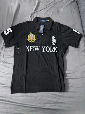 Svart Ralph Lauren New York piké - Säljer en svart pikétröja från Ralph Lauren med stor vit logga och texten NEW YORK på bröstet. Tröjan har broderat emblem i gult och rött, samt nummer 5 på ärmarna. Klassisk krage och knappar framtill. Perfekt för dig som gillar streetstyle.