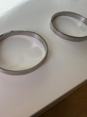 Två silvriga stela armband - Säljer två stilrena silvriga armband i metall. Ett av armbanden har en slät yta medan det andra är dekorerat med små gnistrande stenar längs kanten för extra bling. Perfekta för dig som gillar minimalistisk och modern stil.