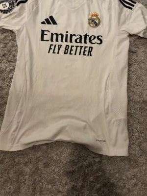 Real Madrid matchtröja Vini Jr. 7 - Säljer en vit Real Madrid matchtröja från Adidas med Vini Jr. och nummer 7 på ryggen. Tröjan har klubbmärke, Adidas-logga och sponsortryck på bröstet samt svarta detaljer på axlarna. Tillverkad i lätt och ventilerande material med AEROREADY-teknologi.