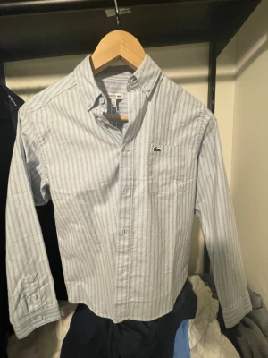 Randig skjorta från Lacoste - Klassisk ljusblå och vit randig skjorta från Lacoste med button-down krage och lång ärm. Skjortan har en bröstficka med den ikoniska krokodilloggan broderad. Tillverkad i mjuk bomull och stängs med knappar framtill.