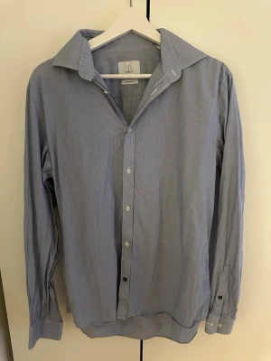 Blåvit randig skjorta från Bläck - Klassisk blåvit randig skjorta från Bläck med lång ärm och knappar framtill. Skjortan har en bred krage och är tillverkad i mjuk bomull, perfekt för en clean och stilren look.