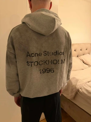 Grå hoodie från Acne Studios - Säljer en grå hoodie från Acne Studios med tryck på ryggen där det står 'Acne Studios STOCKHOLM 1996'. Hoodien har en klassisk huva och ribbade muddar vid ärmslut och nederkant. Perfekt för dig som gillar stilrena och trendiga plagg.