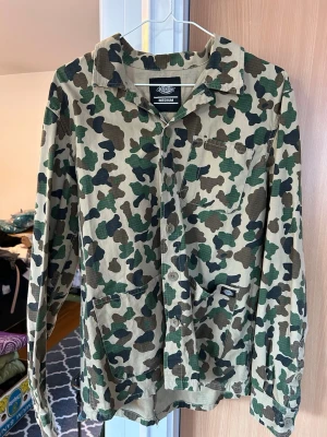 Camouflage jacka från Dickies - Säljer en jacka från Dickies i klassiskt camouflage-mönster. Skjortan har knappar framtill, bröstficka och är gjord i bomull. Perfekt för dig som gillar streetwear och vill sticka ut lite extra.