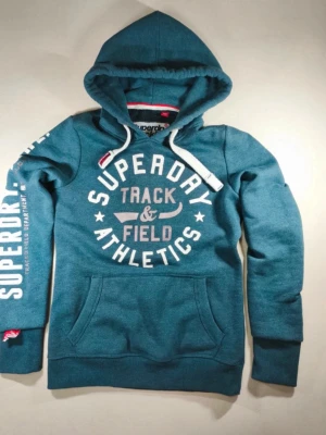 Blå Superdry hoodie med tryck - Snygg hoodie från Superdry med stort vitt tryck på bröstet där det står 'Superdry Track & Field Athletics'. Huva med vita snören och känguruficka framtill. Superdry-logga även på ena ärmen. Perfekt för en sportig och avslappnad stil. Mycket bra skick. Storlek S