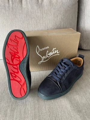 Mörkblå sneakers från Christian Louboutin - Snygga mörkblå sneakers från Christian Louboutin i mocka med klassisk röd sula och diskret logga på sidan. Skorna har rund tå, matchande mörkblå skosnören och beige innersula. Perfekta för dig som vill ha en exklusiv och stilren look. Öppen för byten