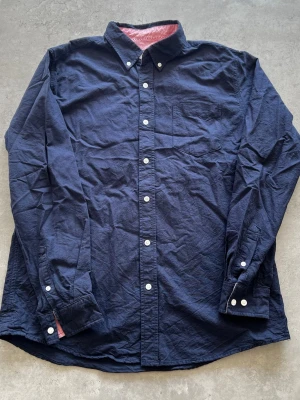 Mörkblå skjorta med vita knappar - L - Stilren mörkblå skjorta med klassisk krage och vita knappar. Skjortan har en bröstficka och långa ärmar med knappslå. Perfekt för dig som gillar enkel och tidlös stil.