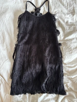 Svart fransklänning  - Säljer en svart kort klänning med smala axelband och massor av fransar som ger en riktigt cool och festlig look. Perfekt för temfest med tex maskeradtema, passar en xs-m