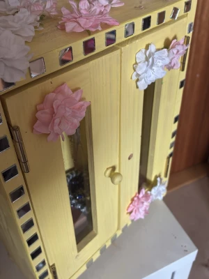 Gult smyckesskåp med glas - Unikt smyckesskåp i trä, målat i gult och dekorerat med spegel runt kanterna. Dörrarna har fönster och är pyntade med rosa och vita konstblommor. Perfekt för att förvara smycken och accessoarer på ett snyggt sätt.
