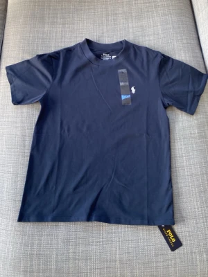 Mörkblå t-shirt från Polo Ralph Lauren - Snygg mörkblå t-shirt från Polo Ralph Lauren med klassisk passform och rund hals. T-shirten har en liten vit logga broderad på bröstet och är tillverkad i mjuk bomull som känns skön mot huden. Perfekt till jeans eller shorts för en clean look.