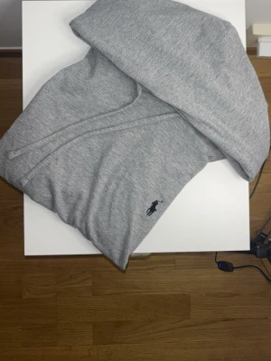 Grå hoodie från Ralph Lauren - Säljer en stilren grå hoodie från Ralph Lauren med klassisk huva och dragsko. Tröjan har det ikoniska svarta Polo-logotypen broderad på bröstet. Tillverkad i mjukt bomullsmaterial som är skönt att bära. Perfekt för en avslappnad och trendig look.