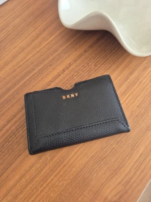 Svart korthållare från DKNY - Snygg svart korthållare från DKNY i äkta läder med guldfärgad logga framtill. Slimmad och stilren design med plats för flera kort. Perfekt att ha i fickan eller väskan när du vill resa lätt och ändå ha koll på dina viktigaste kort. Använt skick!