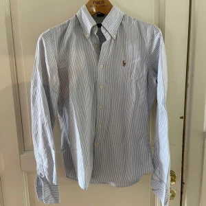 Randig skjorta från Polo Ralph Lauren - Klassisk skjorta från Polo Ralph Lauren med blå och vita ränder. Skjortan har lång ärm, knappar framtill och broderad logga på bröstet. Tillverkad i bomull och har en normal passform. Perfekt för en stilren look.                                                                                                                                                                                                                                                                      obs finns ett par defekter