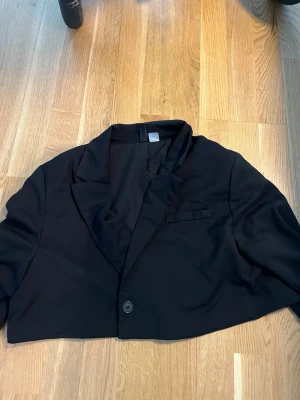 Svart cropped kavaj med knapp - Stilren svart cropped kavaj med en knapp framtill och klassisk krage. Kavajen har lång ärm och en liten bröstficka som detalj. Perfekt för en trendig och modern look. Materialet ser ut att vara en mjuk och lätt tygblandning.