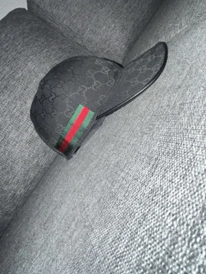 Svart keps från Gucci med GG-mönster - Snygg svart keps från Gucci med det klassiska GG-mönstret över hela kepsen. På sidan finns en grön och röd rand som ger en ikonisk touch. Kepsen har böjd skärm och justerbar rem bak. Perfekt för dig som vill ha en stilren accessoar med lyxig känsla.