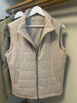 Beige John Henric Väst - Stilren beige dunväst med quiltat mönster och hög krage. Västen har två fickor framtill och stängs med en dragkedja. Perfekt att slänga över en hoodie eller tröja för extra värme. Materialet ser ut att vara en mjuk blandning med dunfyllning.