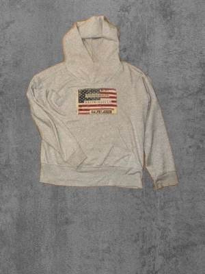 Grå hoodie från Ralph Lauren - Snygg grå hoodie från Ralph Lauren med amerikansk flagga och broderad logga på bröstet. Klassisk huva och ribbade muddar. Perfekt för en avslappnad och cool stil. Tillverkad i mjukt bomullsmaterial.