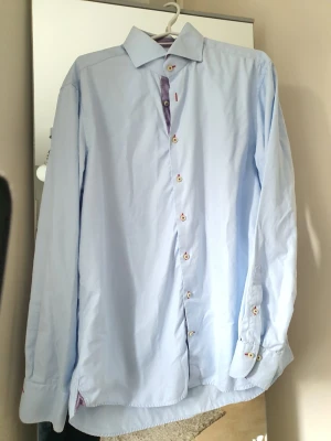 Stenströms skjorta  - Snygg ljusblå skjorta med långa ärmar och klassisk krage. Skjortan har vita knappar med röda knapphål och ett mönstrat tyg i rött och vitt på insidan av knappslån. Perfekt för dig som vill ha en stilren men ändå unik look.