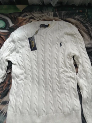 Vit kabelstickad tröja Polo Ralph Lauren - Kabelstickad  vit tröja från Polo Ralph Lauren med klassisk marinblå logga broderad på bröstet. Tröjan har rund halsringning, långa ärmar och ribbade muddar. Perfekt för dig som gillar stilrena och tidlösa plagg.