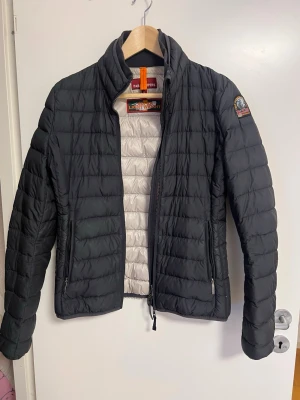 Svart pufferjacka från Parajumpers - Snygg svart pufferjacka från Parajumpers med quiltad design och hög krage. Jackan har dragkedja framtill, två sidofickor med dragkedja och en patch med logga på ärmen. Insidan är ljusgrå och materialet är lätt och smidigt, perfekt för kalla dagar.