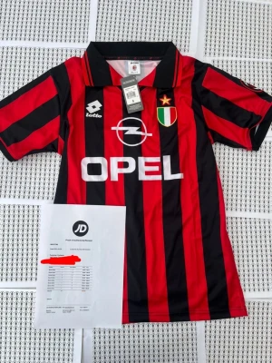 AC Milan retro tröja Maldini #3 - Snygg AC Milan retro matchtröja från Lotto med klassiska röda och svarta ränder. Framsidan har Opel-logga och klubbmärke, baksidan har MALDINI och nummer 3. Kort ärm och krage, perfekt för fotbollsfans. Materialet är lätt och andas.