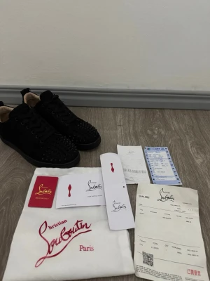 Svarta Christian Louboutin sneakers - Svarta sneakers från Christian Louboutin i mocka med svarta nitar på tån och svarta skosnören. Klassisk låg modell med exklusiv känsla och ikonisk design. Innersulan är ljus och skon har diskreta detaljer som sticker ut.