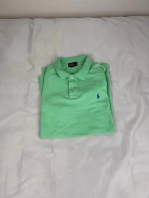 Grön pikétröja från Ralph Lauren - Snygg ljusgrön pikétröja från Ralph Lauren med klassisk krage och tre knappar framtill. Tröjan har korta ärmar och den ikoniska blå loggan broderad på bröstet. Perfekt till jeans eller shorts för en clean look.