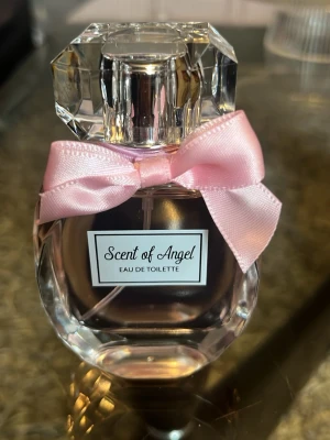 Scent of Angel Eau de Toilette - Elegant parfymflaska i klart glas med en multifacetterad form och en stor, glittrande kork. Flaskan pryds av en söt rosa rosett och etiketten 'Scent of Angel Eau de Toilette' framtill. Perfekt för dig som gillar feminina och lyxiga detaljer.