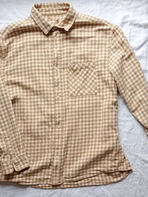 Beige och vit rutig skjorta - Snygg beige och vit rutig skjorta med klassisk krage och bröstficka. Skjortan har långa ärmar och knäppning framtill. Perfekt för dig som gillar en avslappnad men ändå stilren look.