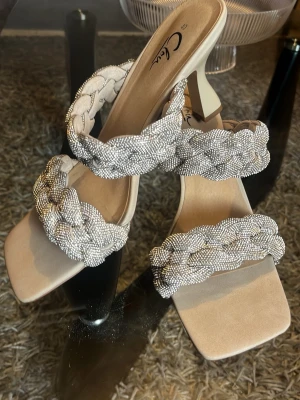 Beige sandaler med flätade pärlband - Säljer ett par beige sandaler från C. Lou med fyrkantig tå och medelklack. Sandalerna har två breda, flätade band täckta av små glittrande pärlor som ger en lyxig känsla. Perfekta för dig som vill sticka ut med snygga detaljer och trendig design.