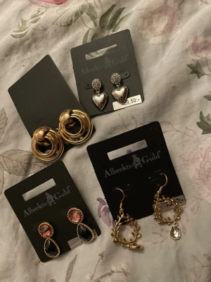 Fyra par örhängen från Albrekts Guld - Säljer fyra par örhängen från Albrekts Guld. Ett par är guldiga ringar, ett par har silverfärgade hjärtan och glittriga stenar, ett par har rosa och svarta droppformade stenar, och ett par är guldfärgade med löv och hängande kristall.