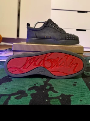 Christian Louboutin svarta sneakers - Svarta sneakers från Christian Louboutin med mattsvart läder och ikoniska röda sulor. Skorna har nitar på tån och klassisk låg siluett. Snygga detaljer och exklusiv känsla, perfekt för dig som vill sticka ut med din stil.