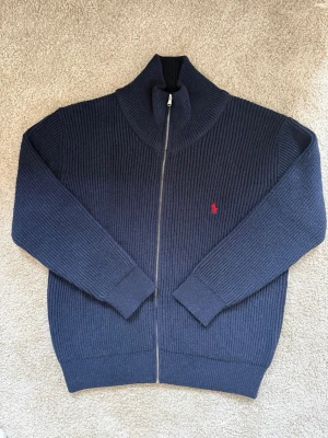 Marinblå stickad ziptröja Polo Ralph Lauren - Mörkblå stickad ziptröja från Polo Ralph Lauren med ribbad struktur och hög krage. Tröjan har dragkedja framtill och den klassiska röda logotypen broderad på bröstet. Strolek XL i mycket bra skick, modellen är 183 cm lång