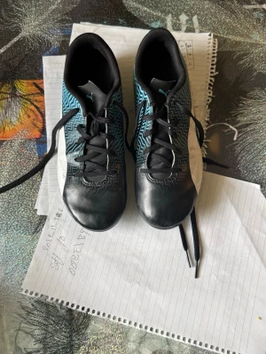 Svarta och blå fotbollsskor från Puma - Säljer ett par snygga fotbollsskor från Puma i svart och blått med vita detaljer och zigzag-mönster på sidorna. Skorna har klassisk snörning och dobbar för bra grepp på planen. Perfekta för dig som vill sticka ut på matchen!