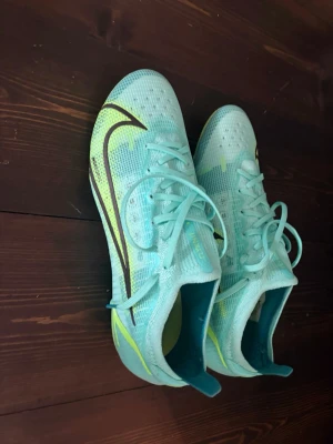 Nike mercurial Vapor 14 elite ag  - Snygga turkosa fotbollsskor från Nike med svart och neongul swoosh på sidan. Skorna har snörning, låg profil och fasta dobbar för spel på gräs. Tillverkade i syntetmaterial som ger bra bollkontroll och passform. Perfekta för dig som vill sticka ut på planen.